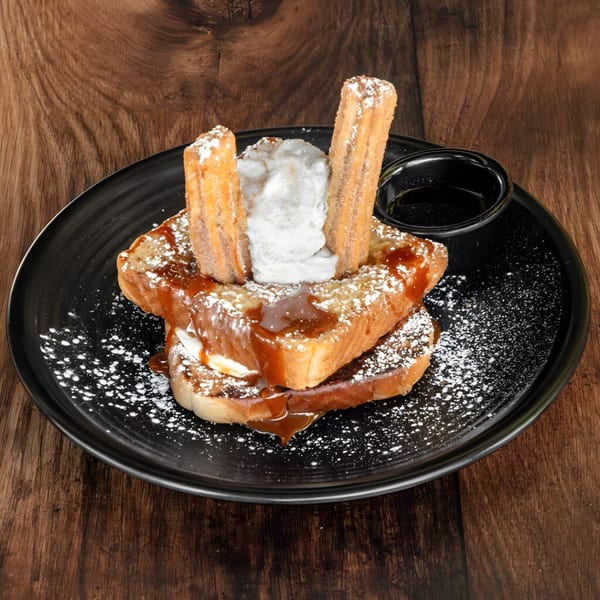 French toast with churros, whipped cream, caramel, and powdered sugar on a black plate.