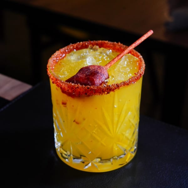 A vibrant yellow Mexican cocktail with a chili-salt rim and a chili pepper garnish.