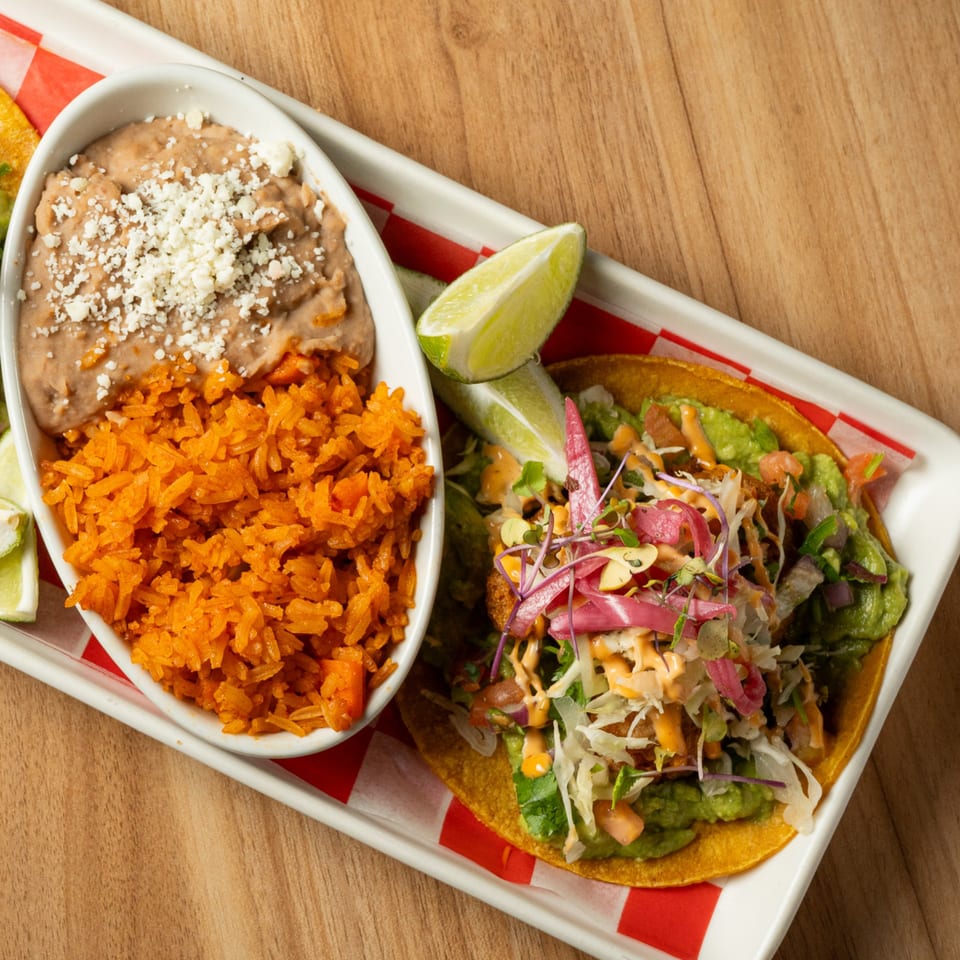 A taco with guacamole, shredded cabbage, and pickled onions, served with rice and refried beans.