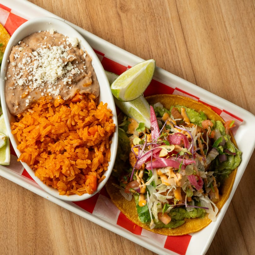 A taco with guacamole, shredded cabbage, and pickled onions, served with rice and refried beans.
