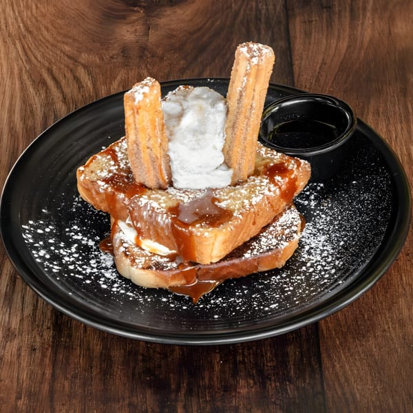 Churro French Toast with whipped cream, powdered sugar, and caramel drizzle on a black plate.