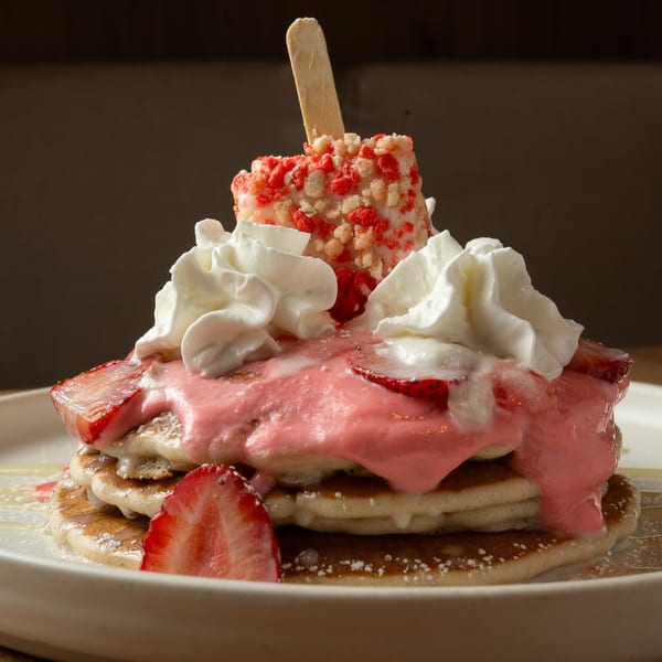 Stack of pancakes topped with pink frosting, whipped cream, strawberries, and a strawberry shortcake bar.