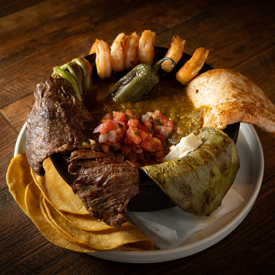 Molcajete with steak, chicken, shrimp, peppers, salsa, and tortillas on a wooden table.
