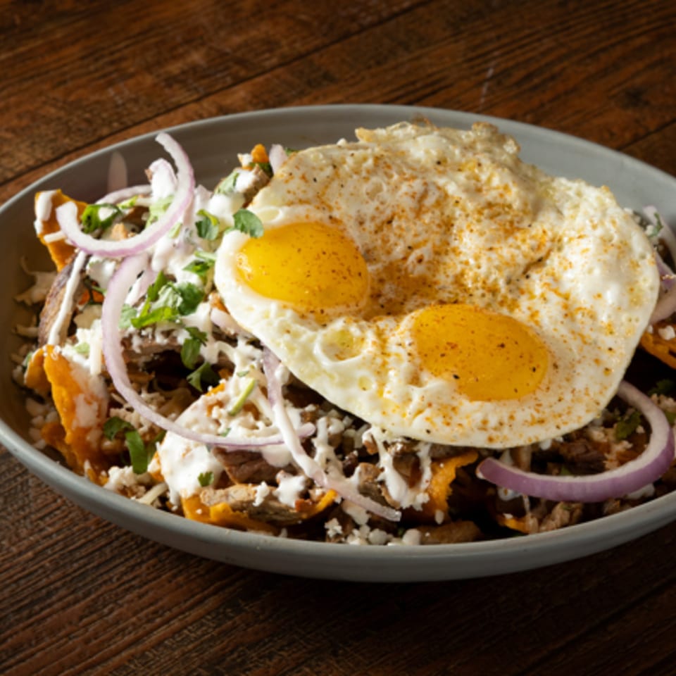 Chilaquiles with two fried eggs, red onion, cilantro, and crema on a gray plate.