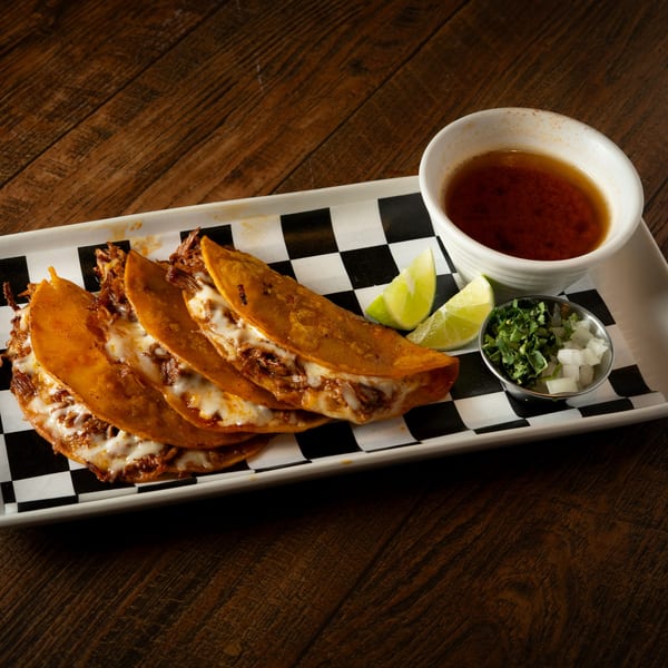 Four cheesy birria tacos with limes, cilantro, onions, and consommé on a checkered paper-lined tray.