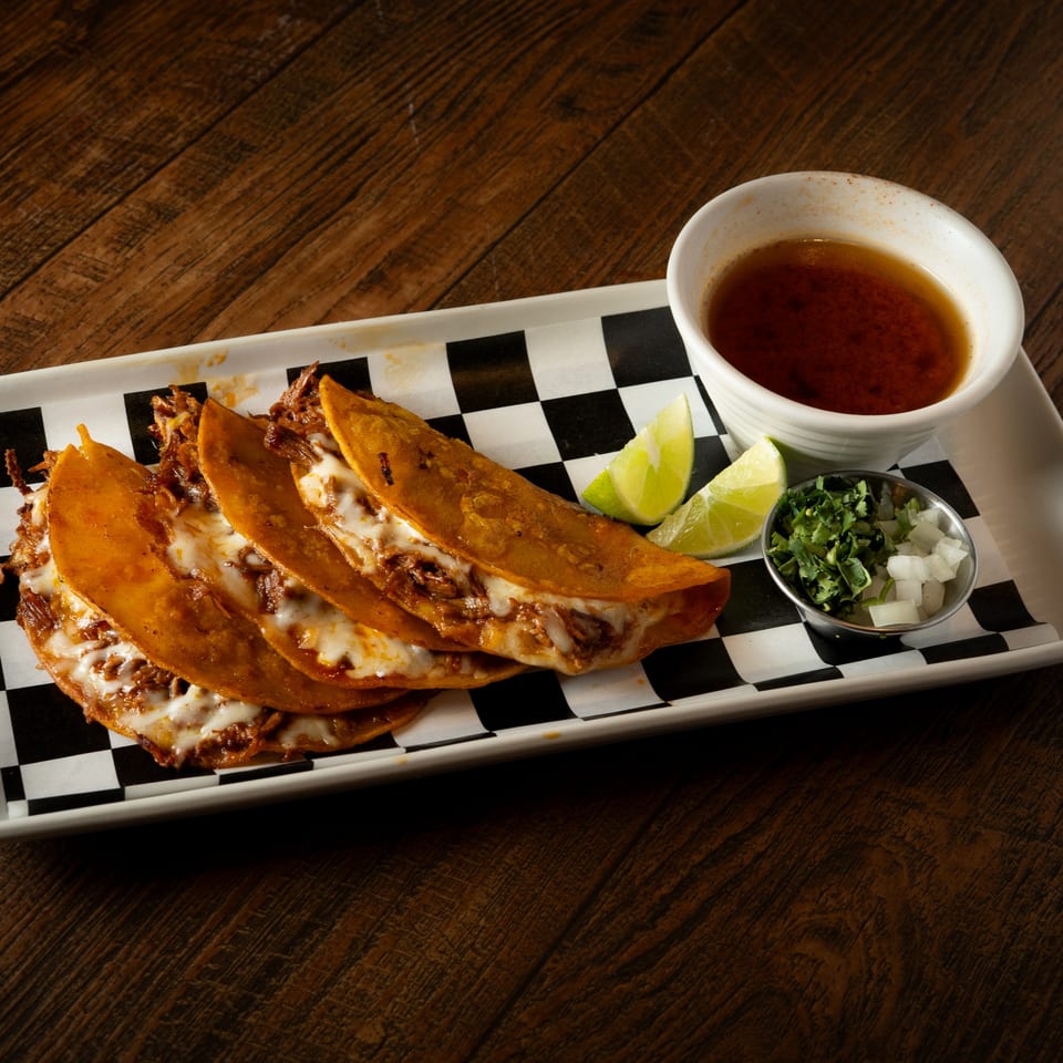 Four cheesy birria tacos with limes, cilantro, onions, and consommé on a checkered paper-lined tray.