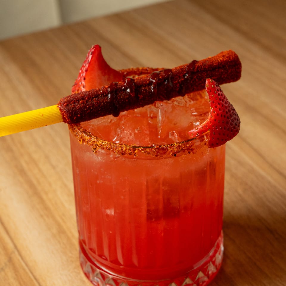 A vibrant red cocktail with ice, garnished with strawberries and a chili-dusted straw.