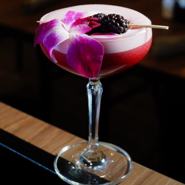 A vibrant pink cocktail in a stemmed glass, garnished with a purple orchid and blackberries.