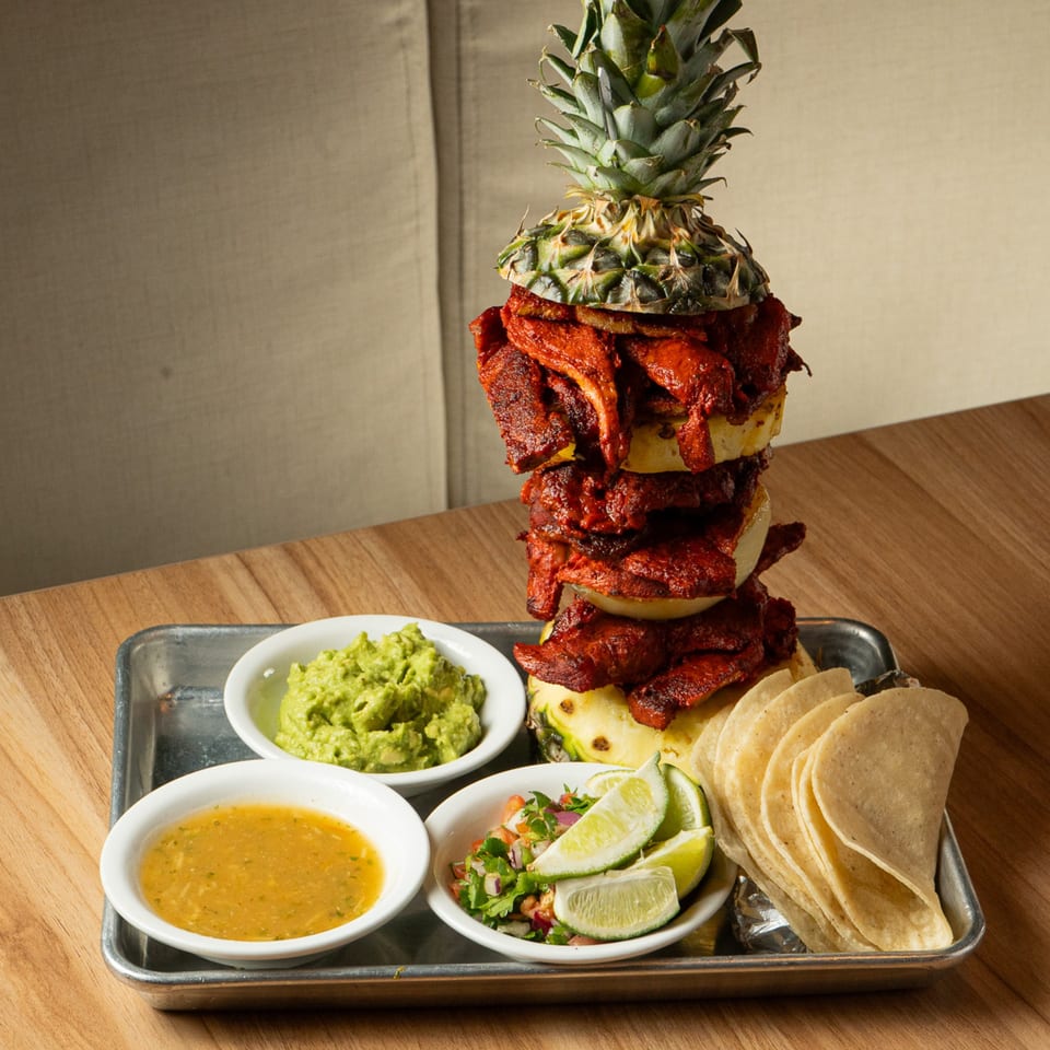 Al pastor meat stacked on pineapple, with guacamole, salsa, pico de gallo, limes, and tortillas.