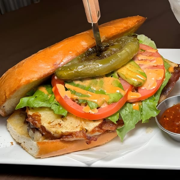 A large Mexican torta sandwich with a knife, filled with lettuce, tomato, avocado, and a green chili.