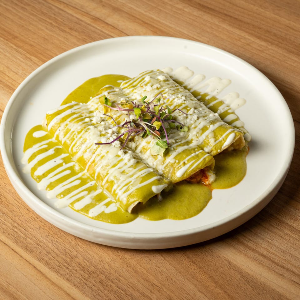 Three green enchiladas covered in green sauce, white crema, crumbled cheese, and microgreens on a white plate.