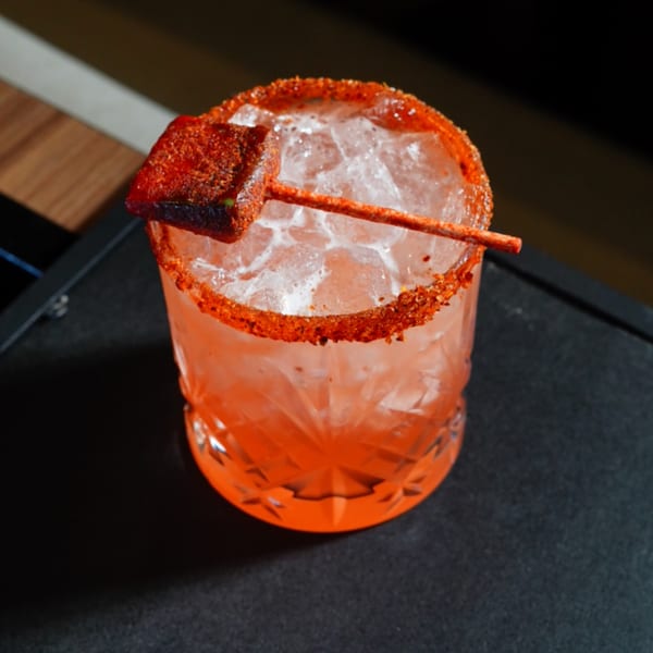 A vibrant red cocktail with a chili-lime rim and a tamarind candy garnish on a dark surface.