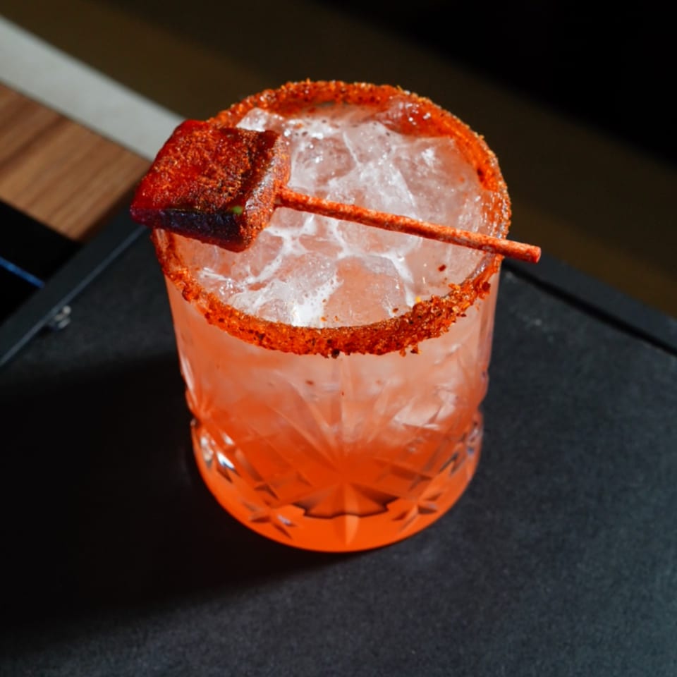 A vibrant red cocktail with a chili-lime rim and a tamarind candy garnish on a dark surface.