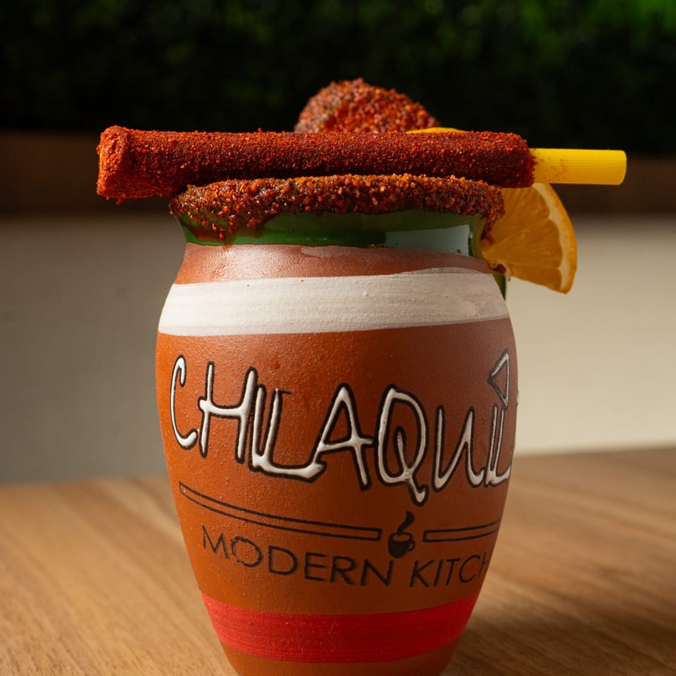 A Chilaquil Modern Kitchen branded clay cup with a drink, garnished with chili-covered candy and an orange slice.