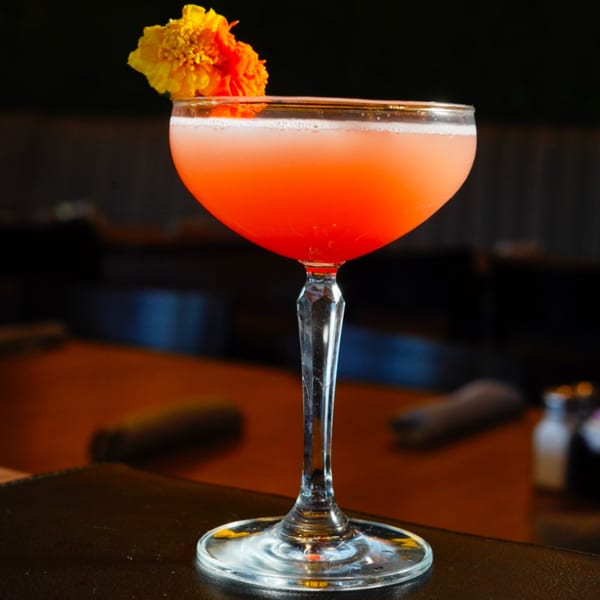 A vibrant orange cocktail with a foamy top, garnished with yellow and orange flowers, on a dark table.