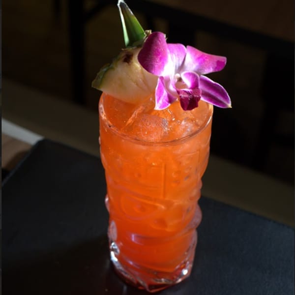 A vibrant orange cocktail in a tiki glass, garnished with a pineapple wedge and a purple orchid.