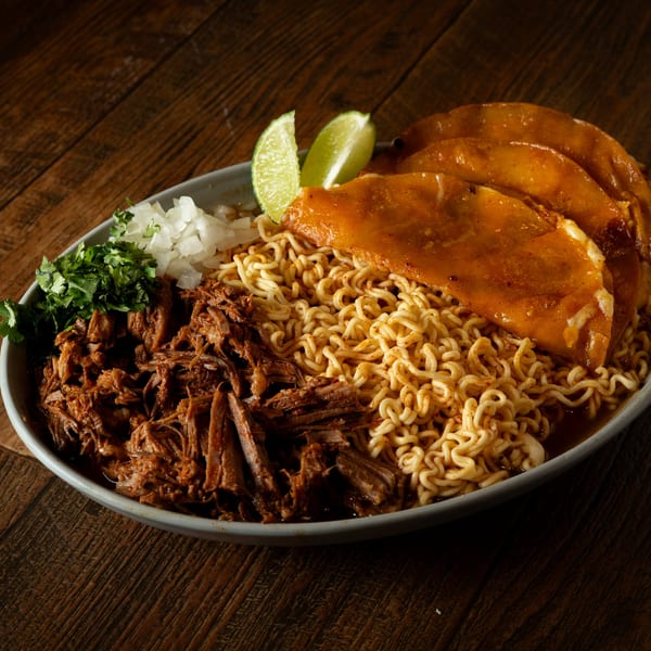 Birria ramen bowl with shredded beef, three birria tacos, cilantro, onions, and lime wedges.