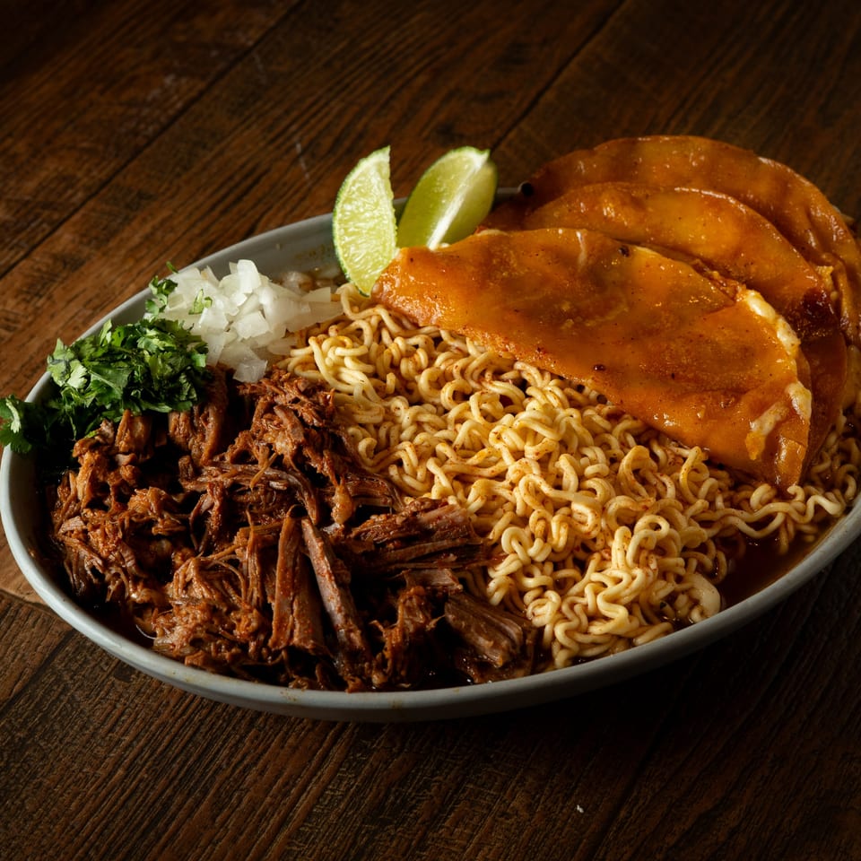 Birria ramen bowl with shredded beef, three birria tacos, cilantro, onions, and lime wedges.