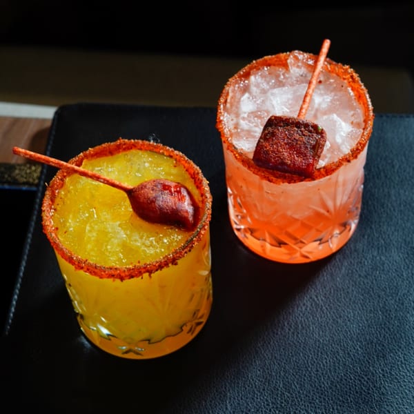 Two vibrant, chili-rimmed cocktails, one yellow and one orange, garnished with chili-dusted candy.